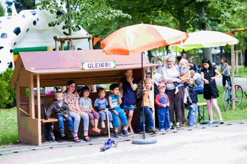 Kinder-Ferien-Stadt im Schützenmattpark – eine Gruppe von Kindern mit Eltern sitzt im Schutz eines Holzhäuschens (Copyright Stefan Bohrer/Robi-Spiel-Aktionen))