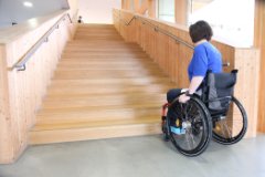 Eine Frau die in einem Rollstuhl sitzt und vor einer langen Holztreppe steht.