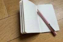 Ein offenes Notizbuch das auf einem Holzboden liegt. Die Seiten sind leer, ein Stift liegt bereit.